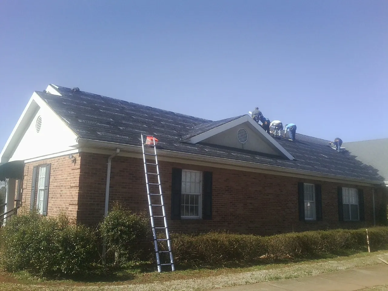 Crew tearing shingles off a brick ranch roof with a ladder set — Metal Roof Repair work by our crew in Semora